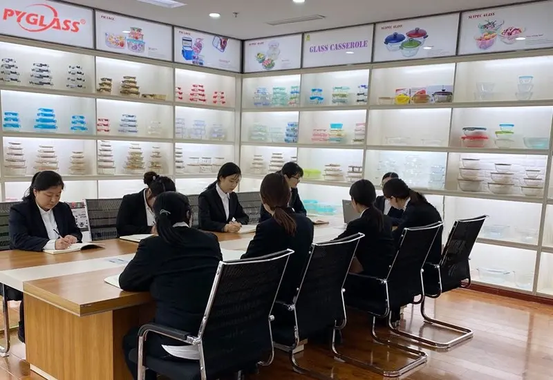 Group of people in formal attire having a meeting in a room with walls of neatly arranged glass kitchen containers on display shelves.