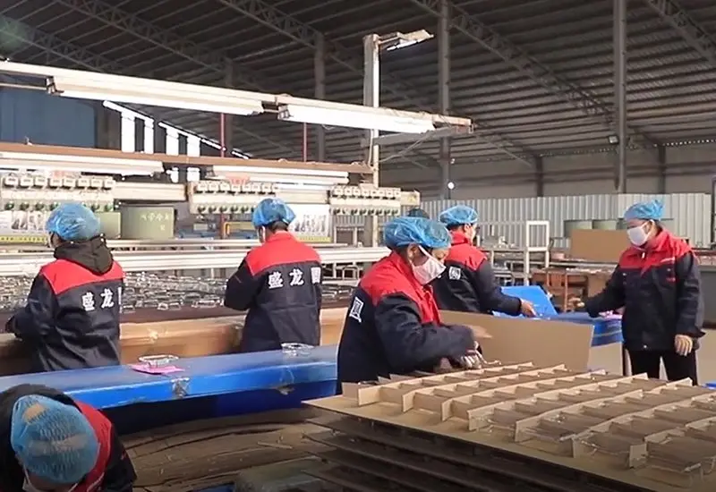 Workers wearing red and blue uniforms assembling and packaging items in a factory setting with machinery in the background.