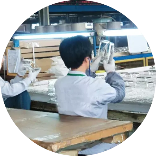 Two workers in a factory inspecting transparent glass containers under bright lights, wearing protective gloves and attire.